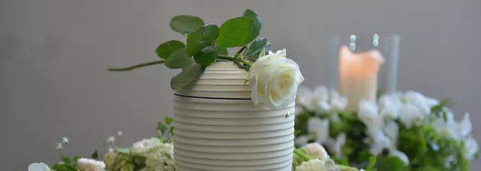 Vase mit einer weißen Rose und grünen Blättern, umgeben von Blumendekoration und einer brennenden Kerze im Hintergrund.