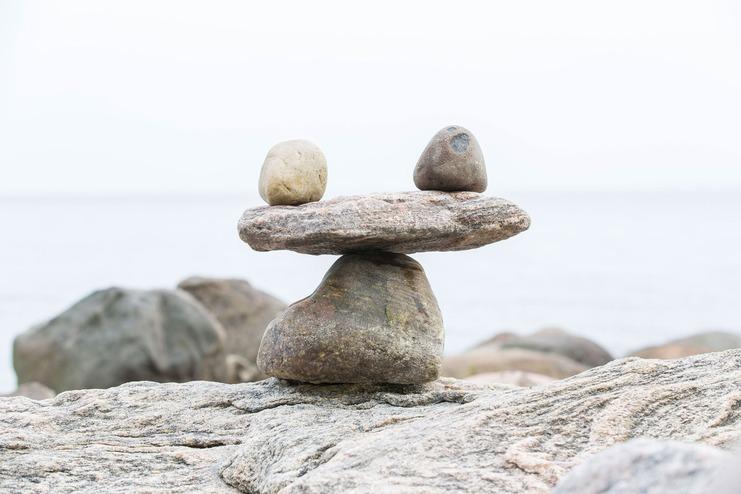 Drei ausgewogene Steine, darunter ein größerer, liegen auf einer felsigen Küste vor einem ruhigen Wasserhorizont.