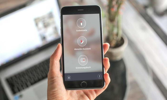 Hand hält ein Smartphone, das eine App mit Funktionen zur Gedenkseite, Abmelde-Assistent und Erinnerungsbuch zeigt.
