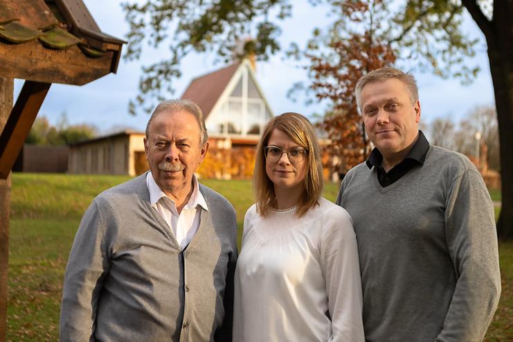 Drei Personen stehen im Freien vor einem modernen Haus, umgeben von Bäumen und herbstlichem Laub.