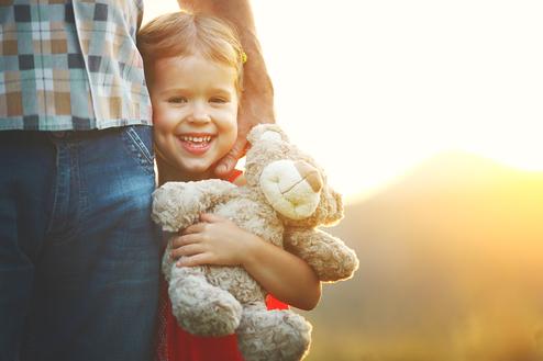 Lächelndes Kind mit einem Plüschteddy steht neben einem Erwachsenen, vor einer sonnigen, verschwommenen Landschaft.